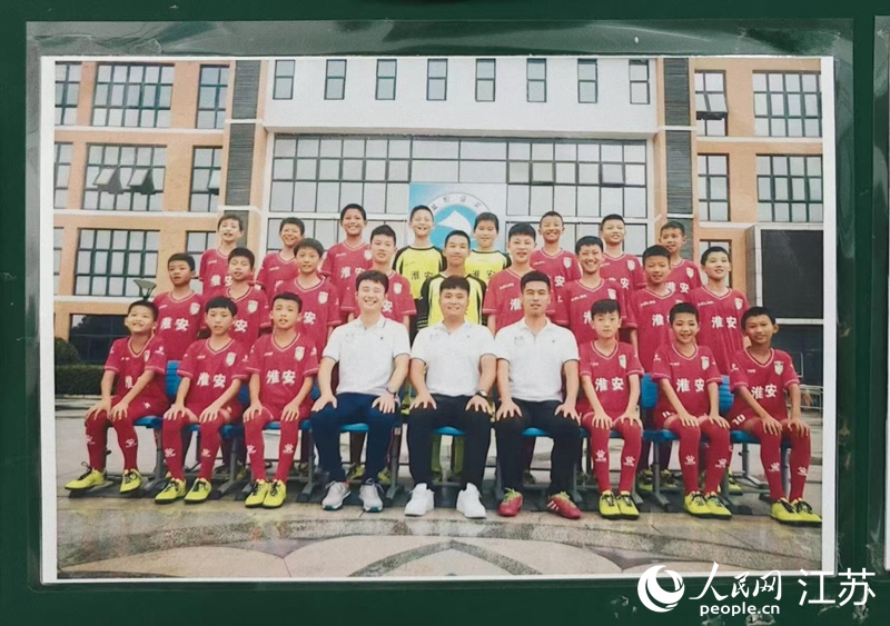 李世恩（上排左四）在淮安工業園區實驗學校05-06組足球隊的合影張貼在教練室墻上，照片中多名球員正效力于“蘇超”淮安隊。 人民網 鞠峰攝
